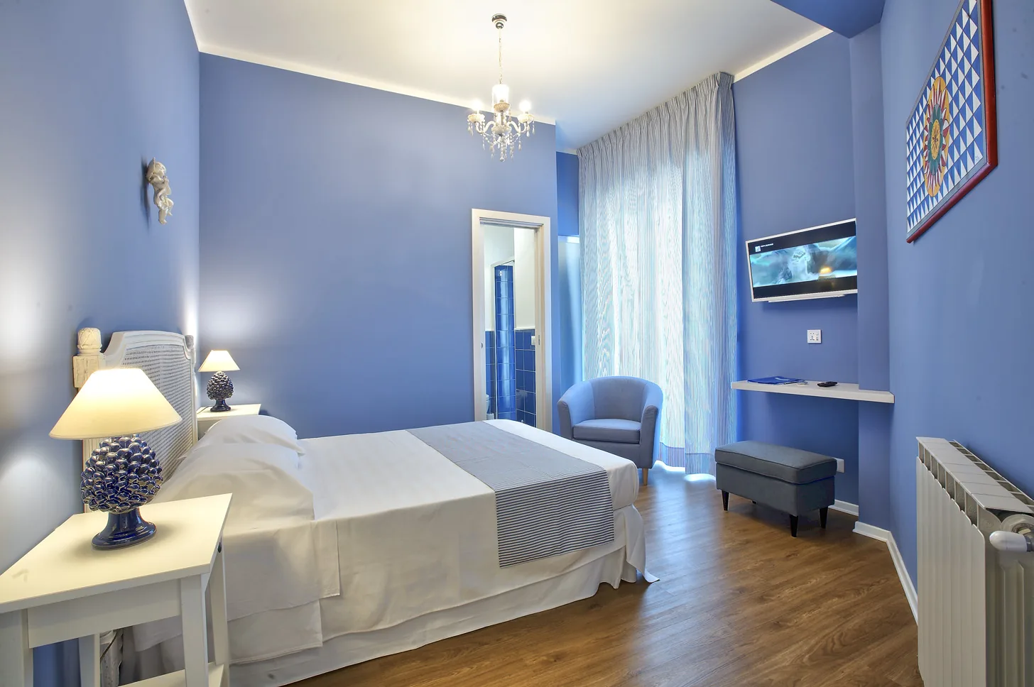 La Camera Blu vista d'insieme: il lampadario di cristallo, la poltroncina alla finestra, la televisione e la porta del bagno