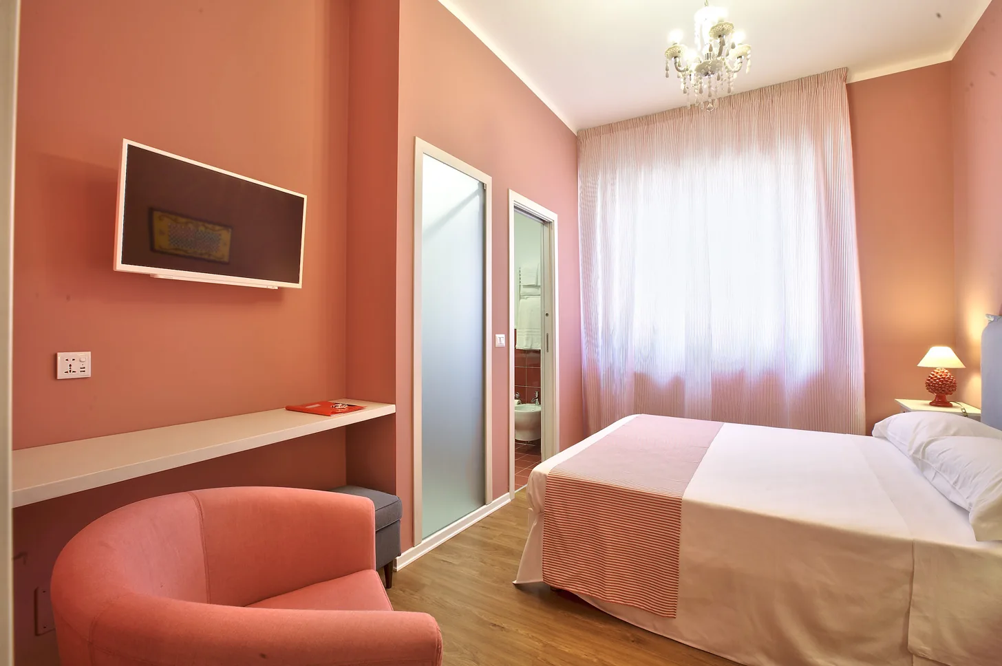 The Red Room from the door: the coral armchair, the shelf-desk, the television and the frosted-glass bathroom door