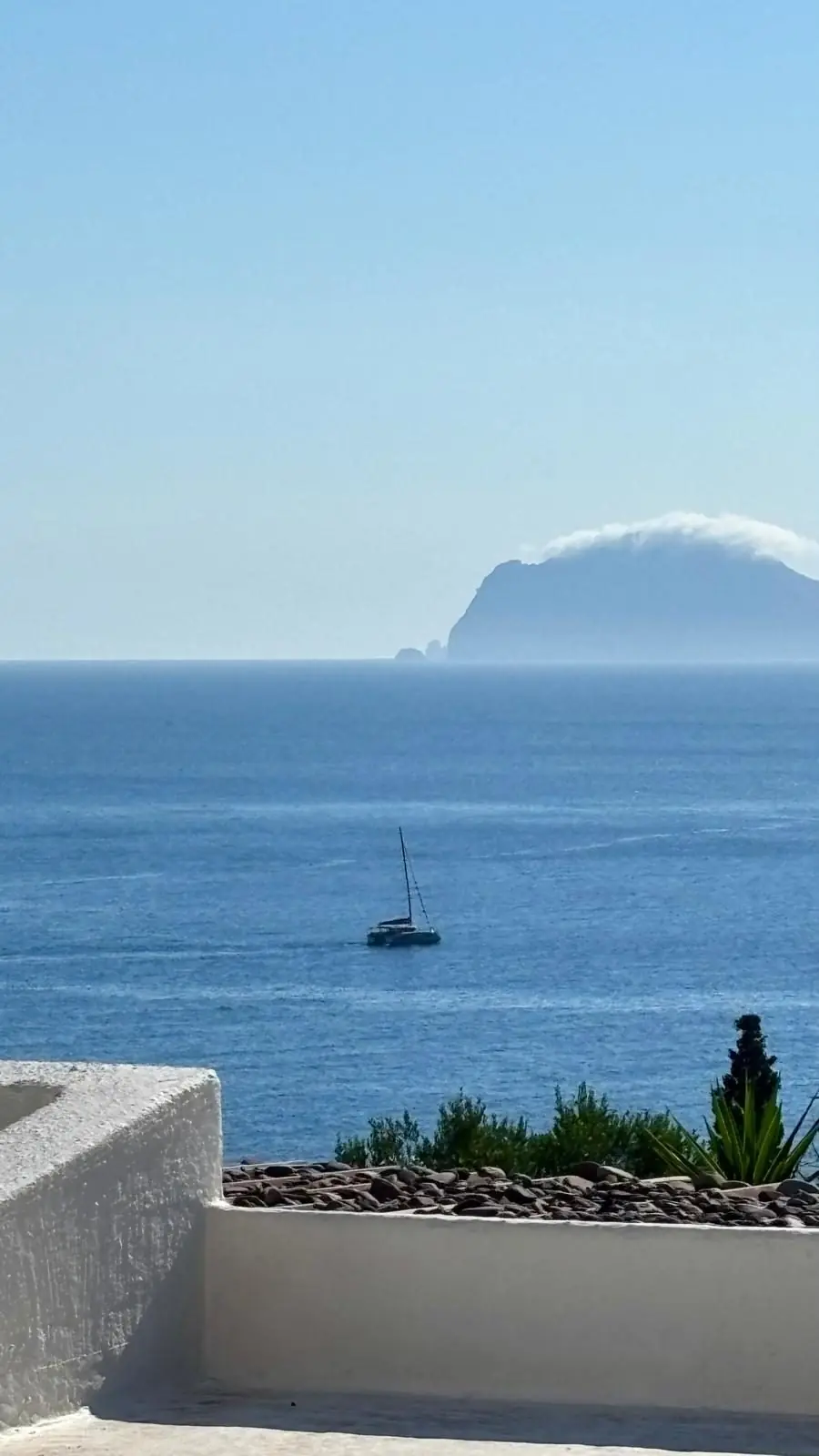 Filicudi under a cap of cloud, a sea stack to its left, seen from a whitewashed terrace on Salina