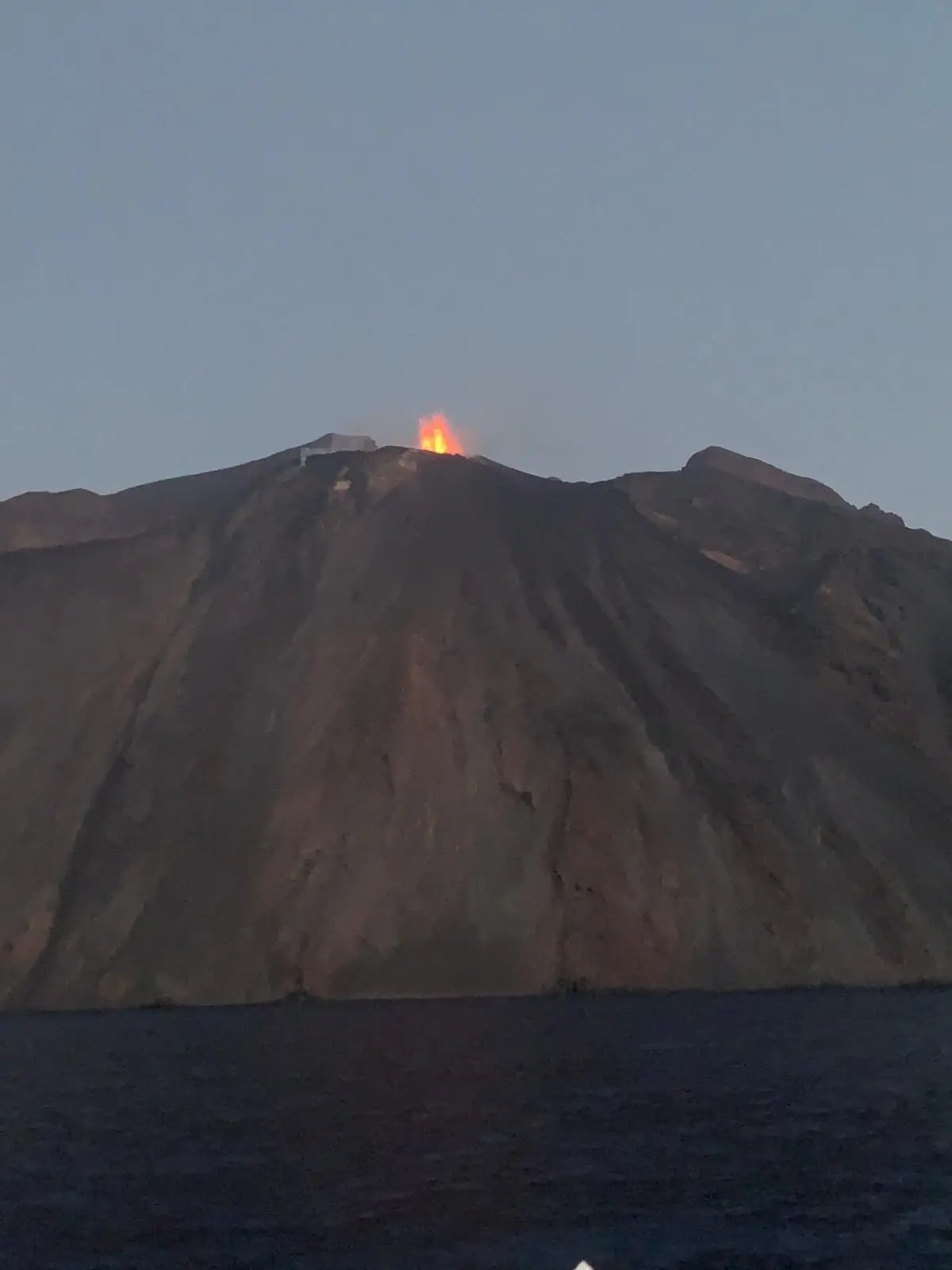 The volcano seen from the sea at dusk, with a red lava fountain rising from the crater